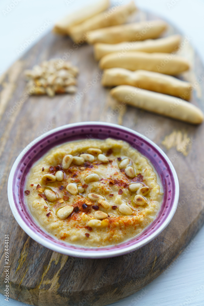 Hummus topped with pine nuts, chili flakes and olive oil, served with bread sticks. Violet bowl, wooden board, high resolution Hummus topped with pine nuts, chili flakes and olive oil, served with bread sticks. Violet bowl, wooden board, high resolution