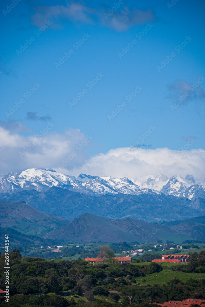 Obraz premium Vertical landscape of valley with houses and with the Picos de Europa snowed in the background in Cantabria, Spain, Europe