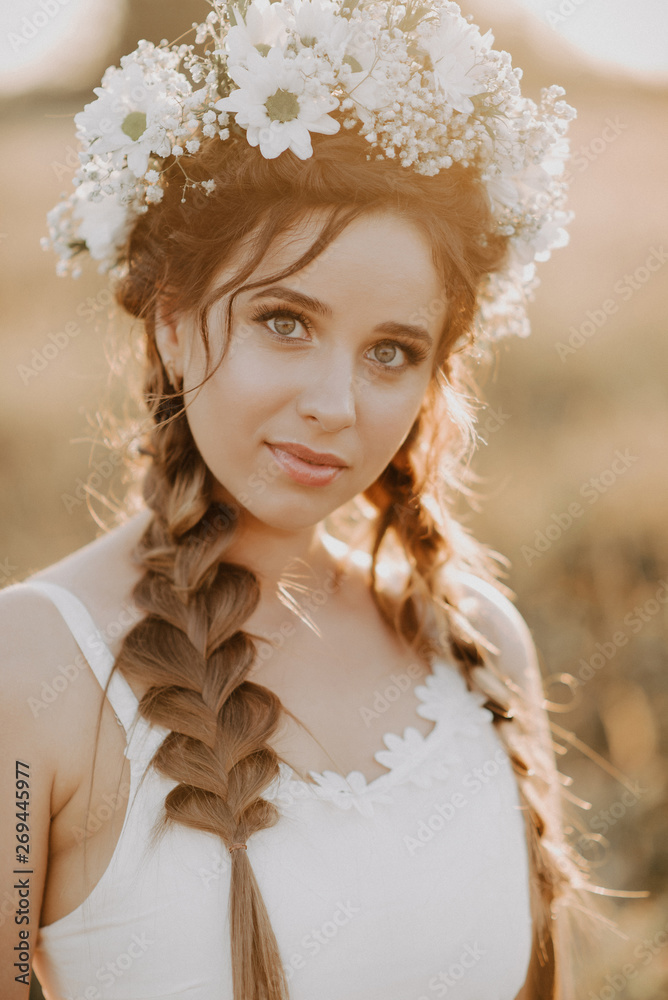 portrait of girl with flower wreath and braids in white dress in summer field at sunset