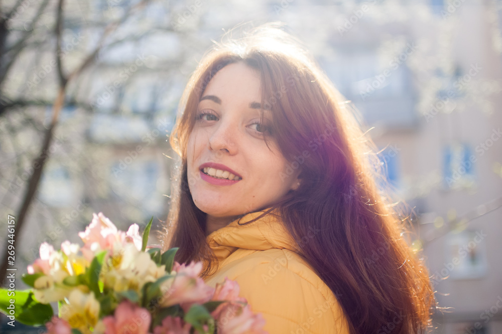 Outdoors female portrait of attractive brunette girl in yellow jacket holding a big bouquet of colorful flowers enjoying spring and looking at tne camera on building background Outdoors female portrait of attractive brunette girl in yellow jacket holding a big bouquet of colorful flowers enjoying spring and looking at tne camera on building background