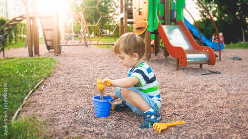 Image of little boy sitting on the playground and puring sand with small plastic spade in colorful bucket. Kid digging and building at park
