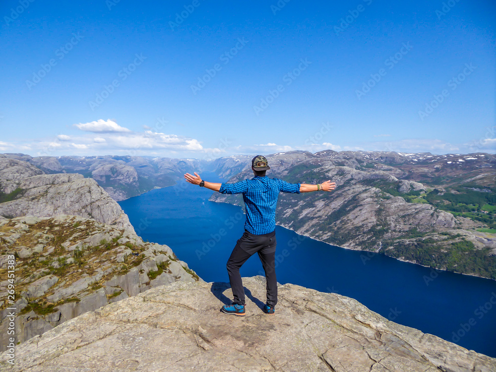 Obraz premium A man wearing blue shirt stands at the edge of a steep cliff of Preikestolen. A view on Lysefjorden. Man enjoys the view, feels free and happy. Man is spreading the arms wide open. Gesture of freedom