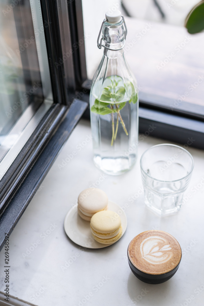 At window of a very cozy summer cafe there is a ficus tree. And near a bottle in which water or lemonade with fresh mint leaves. A cup of hot cappuccino coffee and a saucer of sweet macarons dessert. At window of a very cozy summer cafe there is a ficus tree. And near a bottle in which water or lemonade with fresh mint leaves. A cup of hot cappuccino coffee and a saucer of sweet macarons dessert.