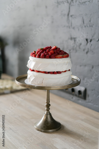 Super delicious summer cream cake dessert with red raspberry berries stands on a vintage stand on a wooden light table against the background of a dark gray wall.