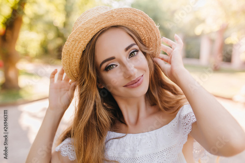 Adorable lady with happy green eyes gently smiling enjoying warm weather in park. Close-up outdoor photo of glad girl holding straw hat with both hands in good mood  walking on the street.