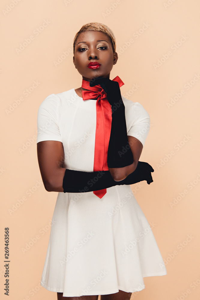 african american woman with short hair posing in white dress, red scarf and black gloves isolated on beige african american woman with short hair posing in white dress, red scarf and black gloves isolated on beige