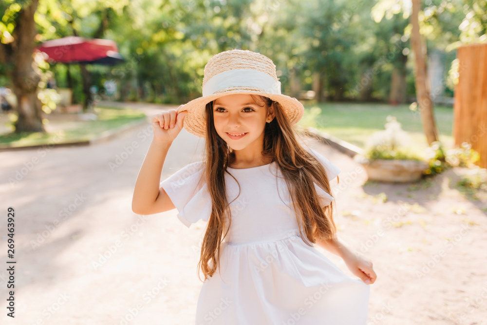 Outdoor portrait of smiling little girl with long straight dark hair walking in park in sunny morning. Cheerful female kid in straw hat and white dress enjoying vacation spending time on the street.