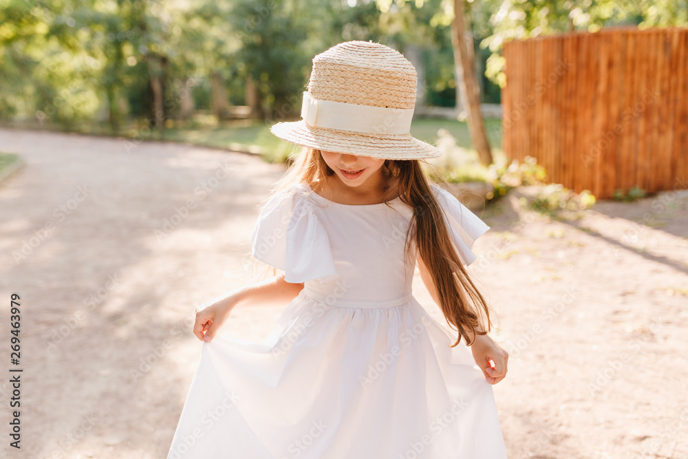 Smiling girl in big straw hat looks at her feet during dance in park. Little lady wears stylish boater playing with white dress enjoying new attire.