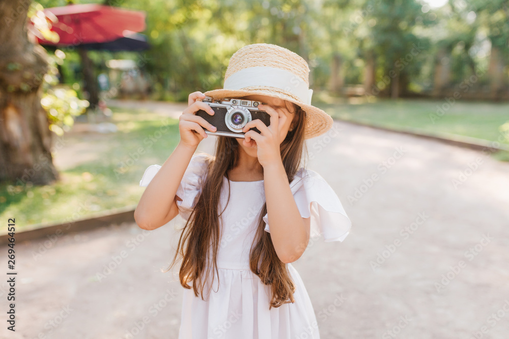 Little girl with long dark hair holding camera in hands standing on the alley in park. Female child in straw hat with white ribbon taking photo of nature view in sunny day.