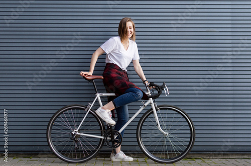 beautiful young girl sit down on a white bicycle on the background of a gray striped wall, the woman is happy