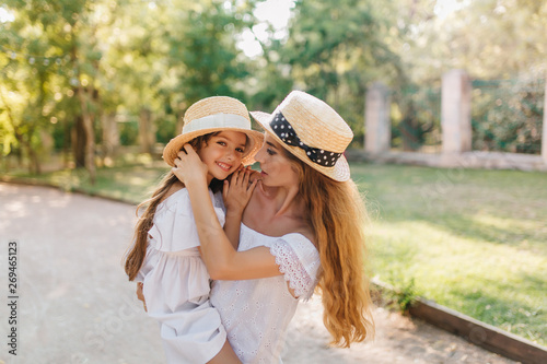 Smiling long-haired girl embracing mother's shoulder and looking to camera. Stylish blonde woman in romantic white attire carrying daughter after walk in park.