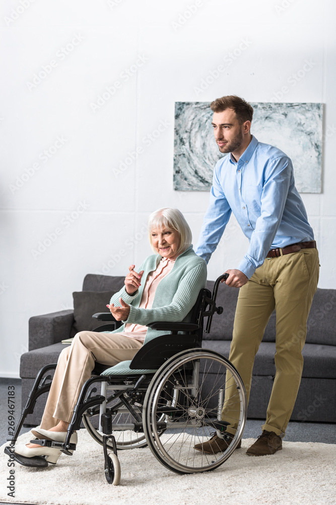 full length view of smiling man carrying disabled senior mother on wheelchair full length view of smiling man carrying disabled senior mother on wheelchair
