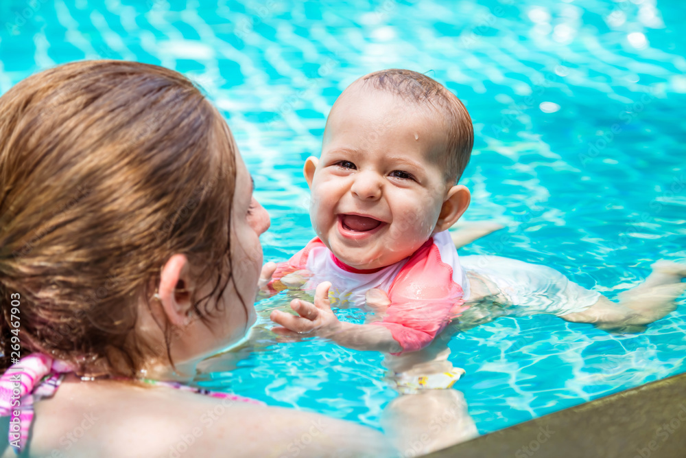 Mother teaches her little baby daughter to swim at the age of 8 months. Summer holiday with infant by the pool at the hotel. Happy laughed children