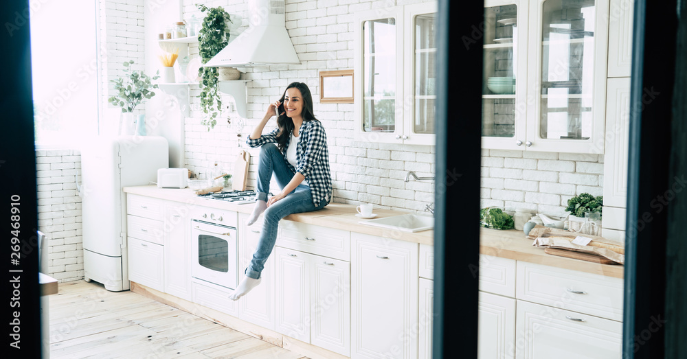 Beautiful cute smiling woman is using smart phone on the kitchen at home. Beautiful cute smiling woman is using smart phone on the kitchen at home.