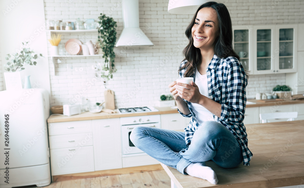 Cute lovely young woman in casual wear with coffee cup in hands is sitting on table in kitchen and relaxing. Cute lovely young woman in casual wear with coffee cup in hands is sitting on table in kitchen and relaxing.