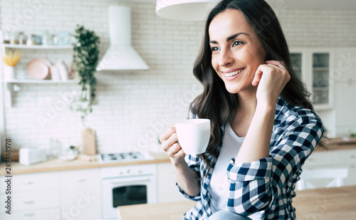 Cute lovely young woman in casual wear with coffee cup in hands is sitting on table in kitchen and relaxing.