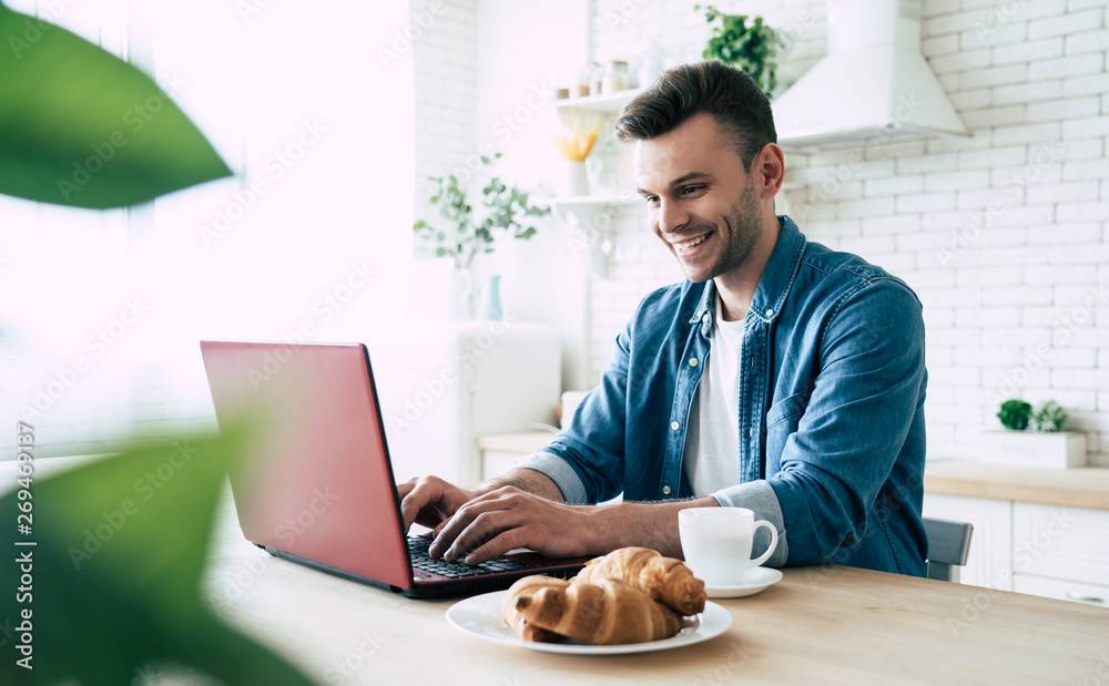 Happy morning. Beautiful smiling man is using laptop and drinks coffee or tea while sitting on the kitchen at home Happy morning. Beautiful smiling man is using laptop and drinks coffee or tea while sitting on the kitchen at home