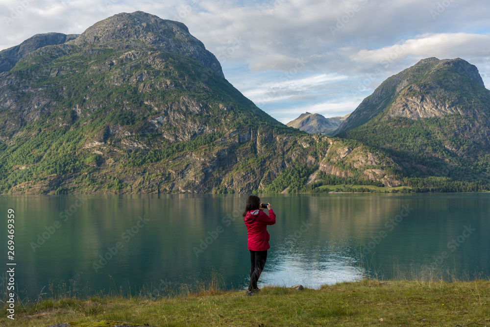 Young caucasian woman in a red jacket taking a photo with her smartphone in a beautiful scene, on the shore of a lake, with a view towards mountains Young caucasian woman in a red jacket taking a photo with her smartphone in a beautiful scene, on the shore of a lake, with a view towards mountains
