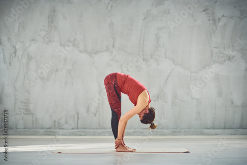 Side view of beautiful Caucasian brunette in red sports wear standing barefoot on the mat in Standing Forward Bend yoga posture. © chika_milan