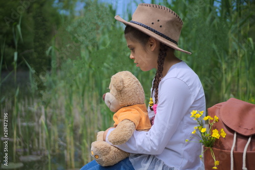 a beautiful little girl by the river sits and enjoys with her toy bear