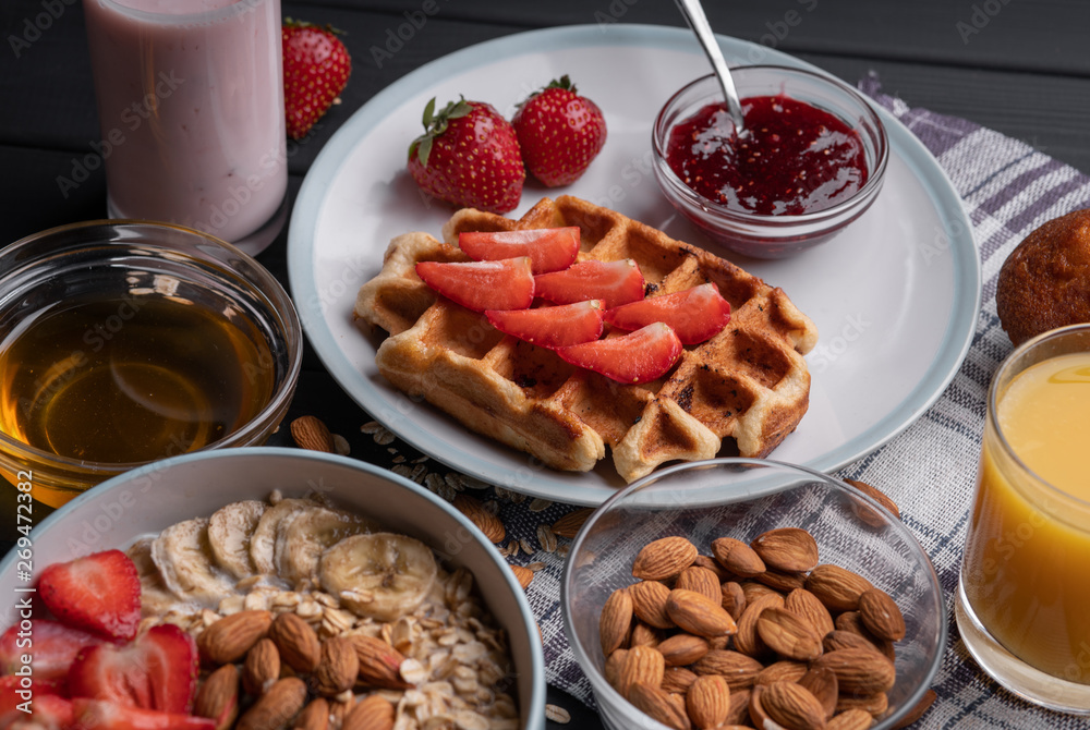 Breakfast. Healthy food - milk oatmeal with a belgian waffle with fruit, honey, strawberry jam, chocolate muffin and fresh yogurt. Set meal on black table background. Top view Breakfast. Healthy food - milk oatmeal with a belgian waffle with fruit, honey, strawberry jam, chocolate muffin and fresh yogurt. Set meal on black table background. Top view