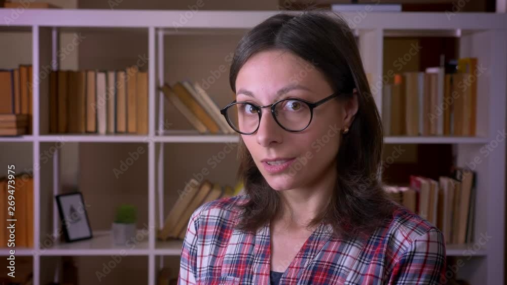 Closeup shoot of young attractive female student in glasses smiling with shyness looking at camera in the library indoors Closeup shoot of young attractive female student in glasses smiling with shyness looking at camera in the library indoors