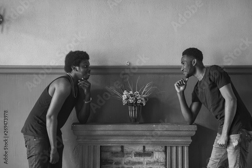 Two handsome adult african male friends in casual clothing standing near deocrative fireplace and looking at each other. Couple of african american men staring at each other in front of brown wall. photo