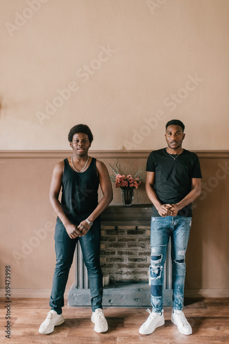 Two handsome adult african male friends in casual clothing posing together like gangsters in front of decorative fireplace on wall. Two confident african american men indoor lifestyle portrait.