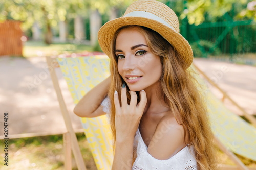 Close-up portrait of inspired girl with lightly tanned skin playing with her long golder hair. Outdoor photo of smiling young woman in vintage boater and white summer dress.
