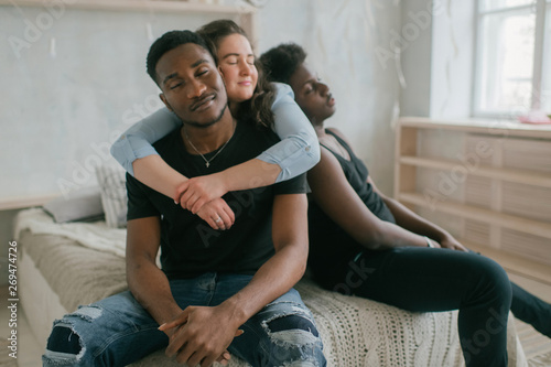 Multicultural love and relationships concept. Young white woman sits and hugs with two african dark skinned men. Soft focus studio portrait of interracial embracing couple. Interracial loving trios.