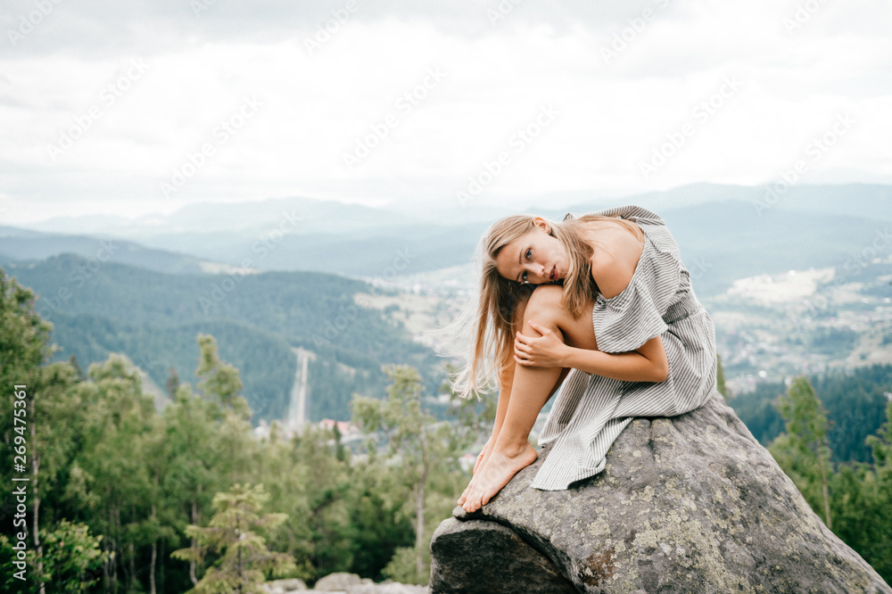 Naklejka premium Lonely young girl sits at stone on top of mountain with far view at hills and cloudy sky and hugs her legs with head lying on knees. Beautiful blonde woman outdoor summer portrait. Female at nature.