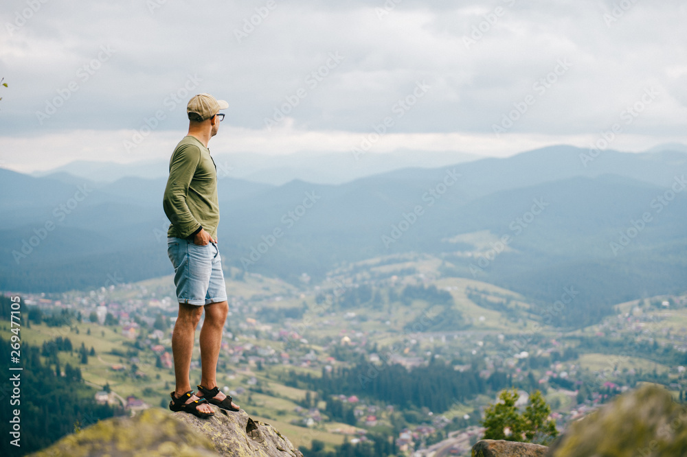 Lifestyle summer portrait of successful man standing on top of mountaing with beautiful landscape in front. Male traveler enjoying nature view from highest peak at hill.