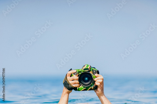 Male diver swimming under water and keeping dry and safety professional photocamera at his hand above water in ocean. Crazy photographer making photos from deep sea. Funny and dangerous hobby and job
