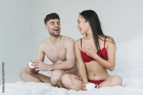 cheerful interracial couple talking while sitting on bed with crossed legs and holding coffee cups