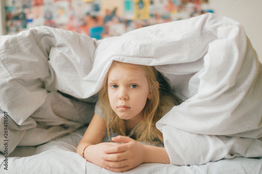 Young adorable blonde girl with funny face lying on bed under white blanket and look at camera with sad eyes. Little female child with pensive face indoor lifestyle portrait. Little girl portrait. Young adorable blonde girl with funny face lying on bed under white blanket and look at camera with sad eyes. Little female child with pensive face indoor lifestyle portrait. Little girl portrait.