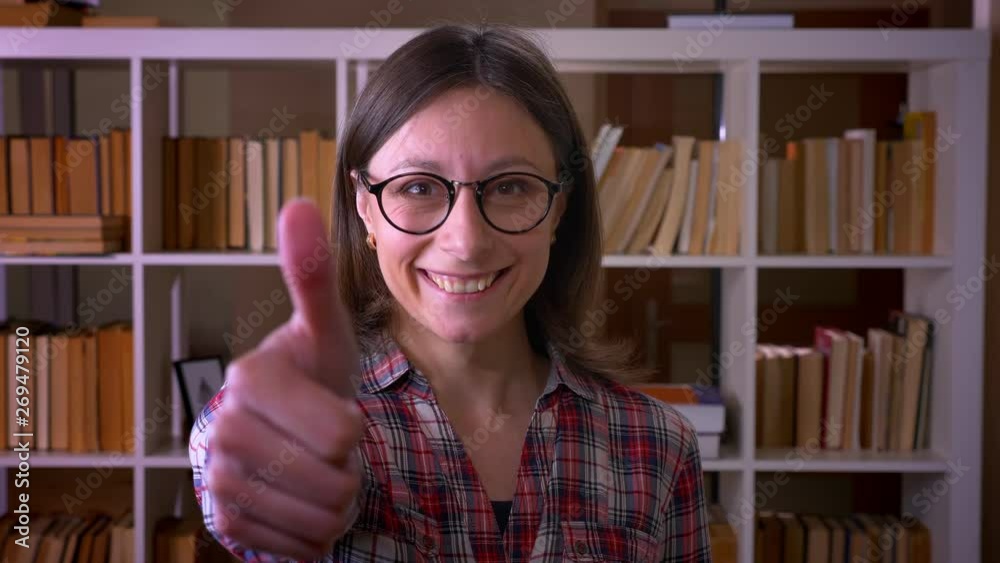 Closeup shoot of young attractive female student in glasses showing a thumb up looking at camera in the university library indoors Closeup shoot of young attractive female student in glasses showing a thumb up looking at camera in the university library indoors
