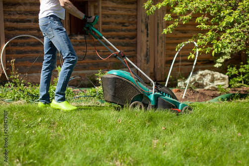 Young man mowing the lawn. Worker doing his job in backyard. Spending summer day in garden. Lawnmower standing on the background of private garden. Close up..