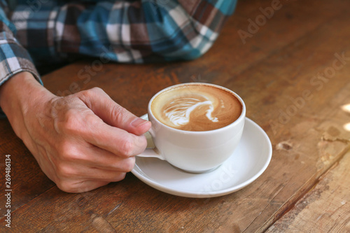 Man in casual blue chess shirt was holding a white cup of hot cappuccino with white foam for drinking in the morning in rustic style.