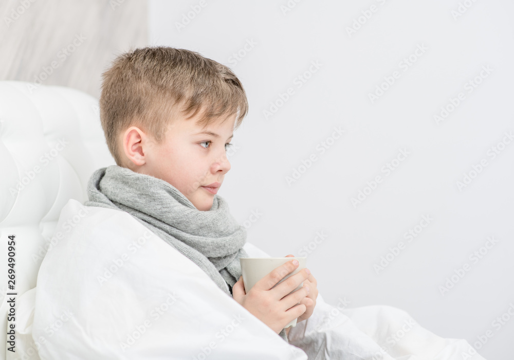 Sick boy with warm scarf sitting on the bed with cup. Empty space for text Sick boy with warm scarf sitting on the bed with cup. Empty space for text