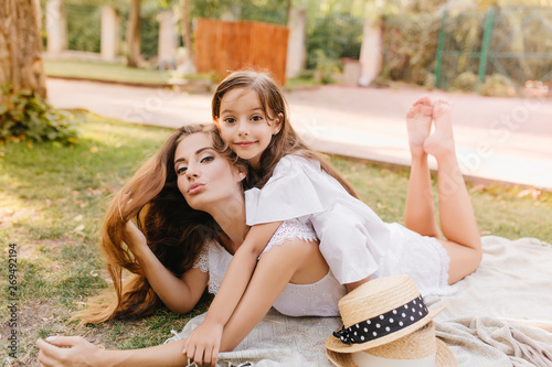 Obraz na plátne Attractive tanned woman with kissing face expression lies on blanket in sunny park playing with kid
