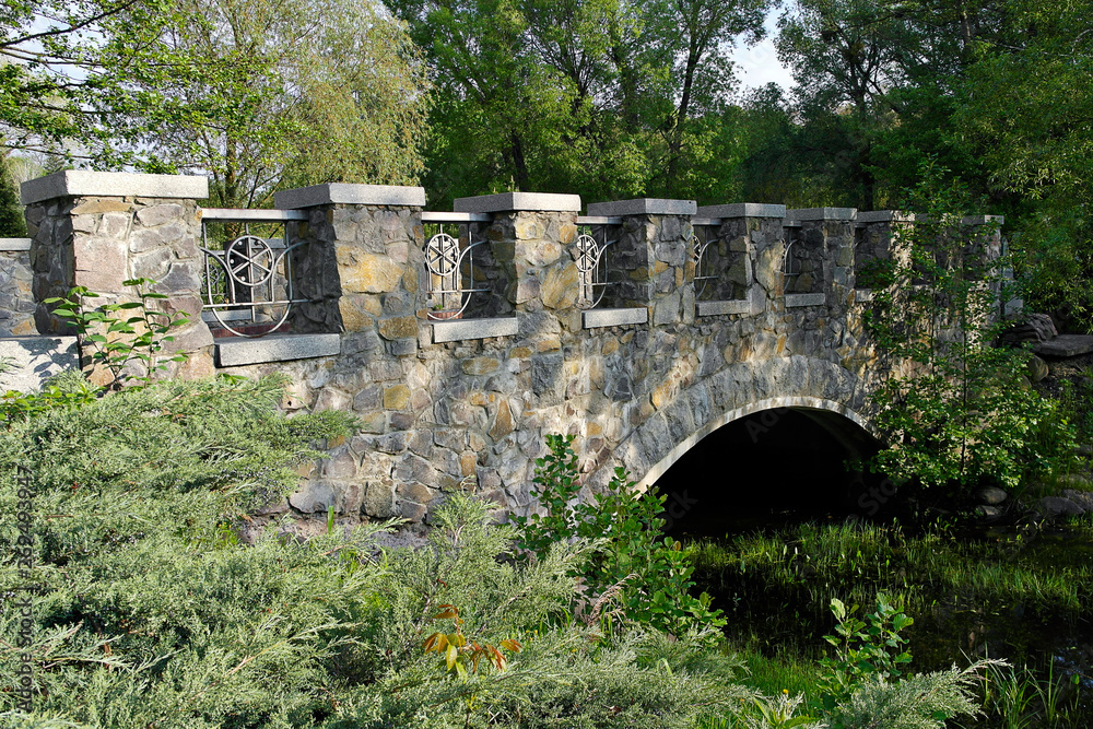 Stone bridge over the creek. flowing among large green trees and bushes. Early summer morning.