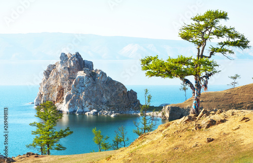 Fototapeta Naklejka Na Ścianę i Meble -  Baikal Lake. Olkhon Island on a summer day. The famous Burkhan Сape, the magic Shamanka Rock and the Wish larch tree with colorful ribbons of tourists in the sunset light