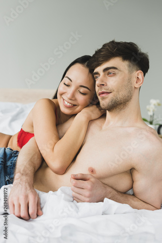 happy asian woman embracing pensive shirtless boyfriend in bedroom