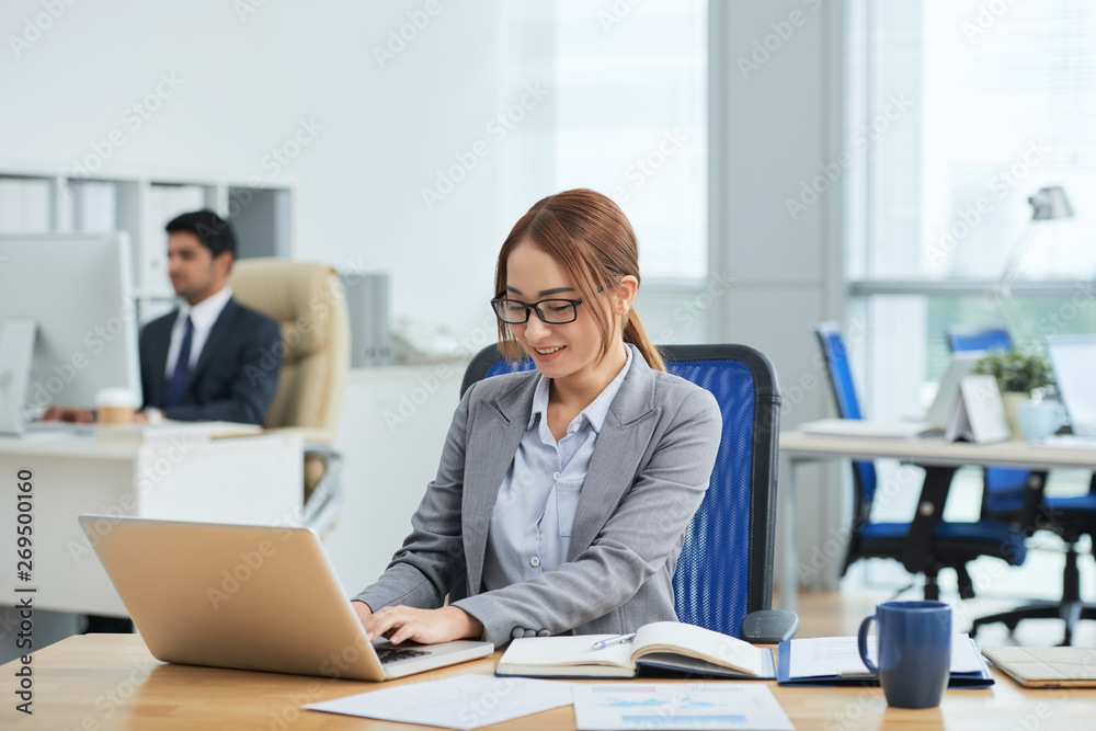 Smiling young secretary in eyeglasses sitting at the table and typing on laptop computer at office with businessman working in the background Smiling young secretary in eyeglasses sitting at the table and typing on laptop computer at office with businessman working in the background