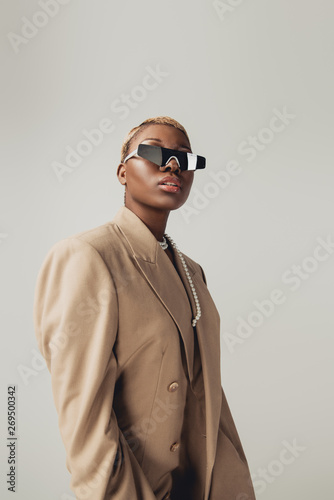 fashionable african american girl posing in sunglasses and beige jacket isolated on grey