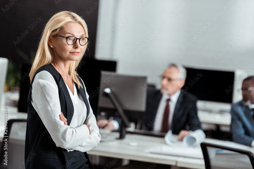 selective focus of attractive businesswoman in glasses standing with crossed arms near multicultural men selective focus of attractive businesswoman in glasses standing with crossed arms near multicultural men