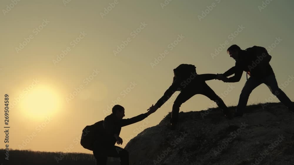 Silhouettes of tourists climbing mountain cliffs against the backdrop of a sunset, helping each other's hand. Help in the mountains and teamwork in hiking. Teamwork concept.