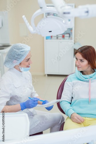 Young beautiful female dentist is talking to her patient. Red-haired woman smiling at the reception at the doctor. Dental services.