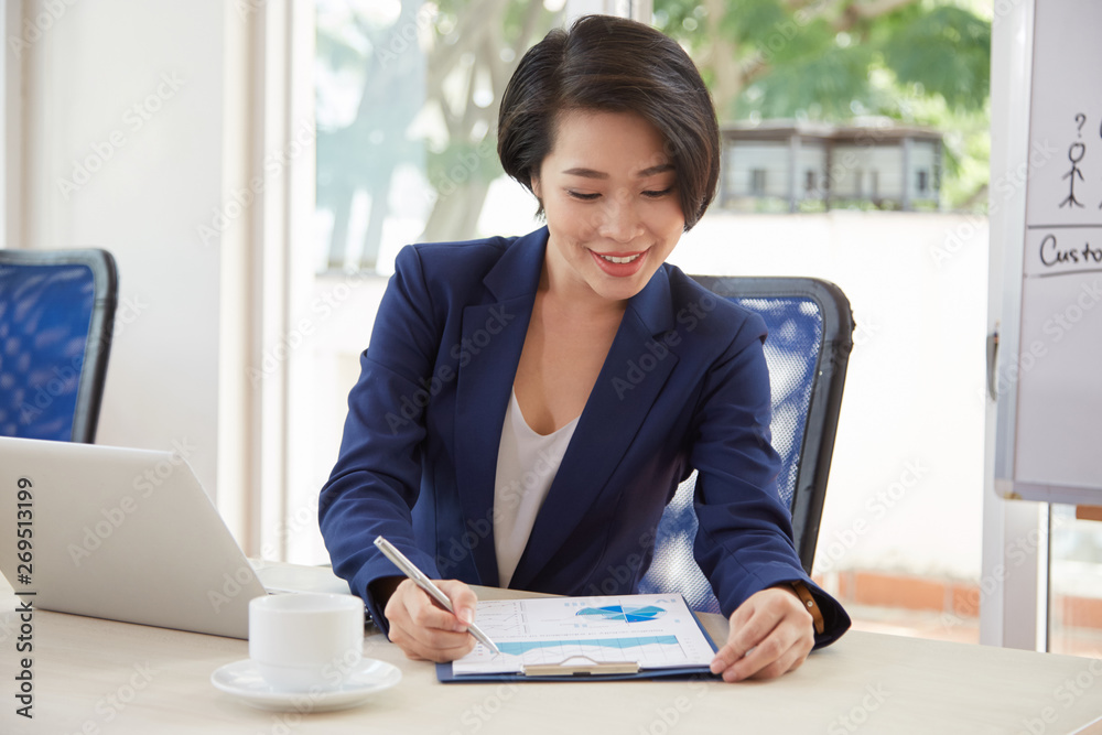 Asian young businesswoman sitting at workplace with laptop examining financial graphs and smiling while working at office Asian young businesswoman sitting at workplace with laptop examining financial graphs and smiling while working at office