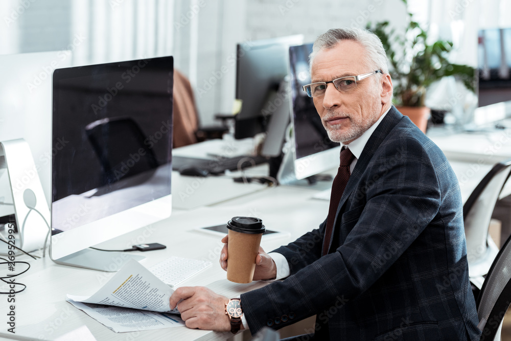handsome mature businessman holding newspaper and paper cup in office